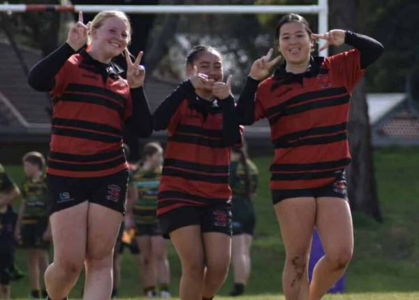 Three girls in Onkaparinga Rugby Club uniforms smile for the camera, giving peace signs and thumbs up.