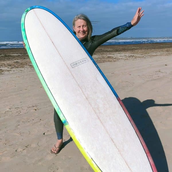 Sue, a participant in the SMART Recovery program, smiling behind a surfboard on a beach.